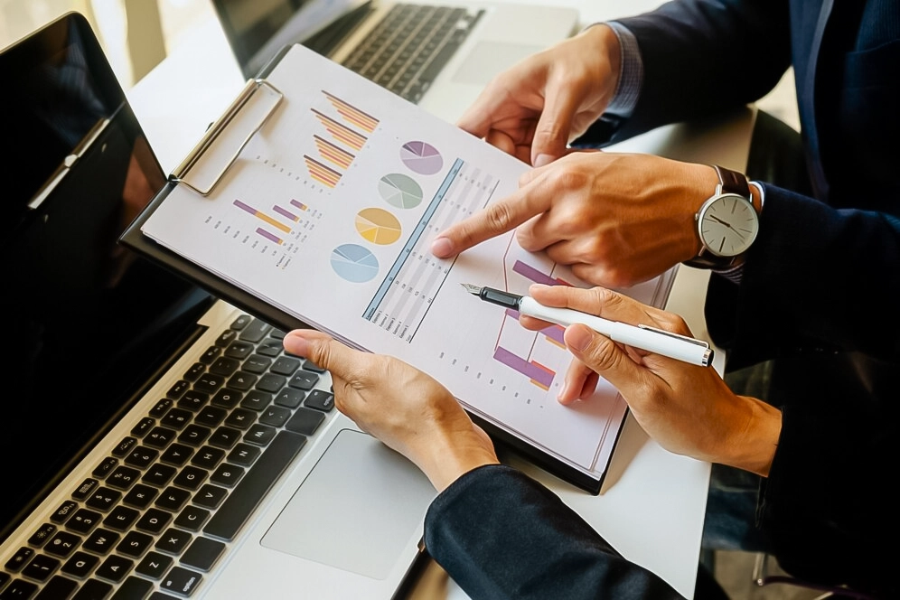 Dois homens em frente ao computador com gráficos e calculadora